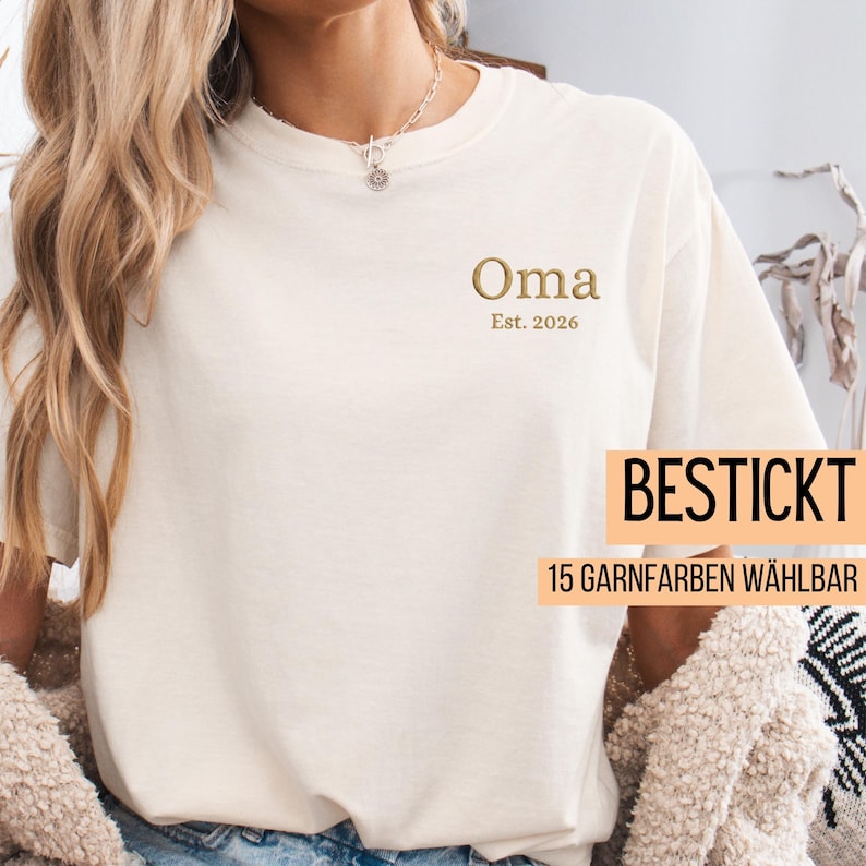 May include: Cream-colored t-shirt with the word "Oma" embroidered in gold thread, along with "Est. 2026". The shirt also features the text "BESTICKT" and "15 GARNFARBEN W&Auml;HLBAR" in a rectangular box.
