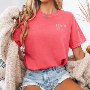 May include: A coral-colored t-shirt with the word "Oma" and the text "Est. 2026" in a light color. The shirt is short-sleeved and has a crew neck. The person is wearing denim shorts and a cream-colored cardigan.