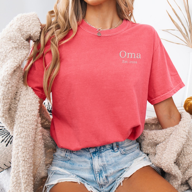 May include: A coral-colored t-shirt with the word "Oma" and the text "Est. 2026" in a light color. The shirt is short-sleeved and has a crew neck. The person is wearing denim shorts and a cream-colored cardigan.