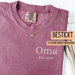 May include: A dusty rose t-shirt with the word "Oma" embroidered in white, along with "Est. 2026". A gold necklace with a pendant rests on the shirt. The shirt has a Comfort Colors tag and text that reads "BESTICKT" and "15 GARNFARBEN W&Auml;HLBAR".