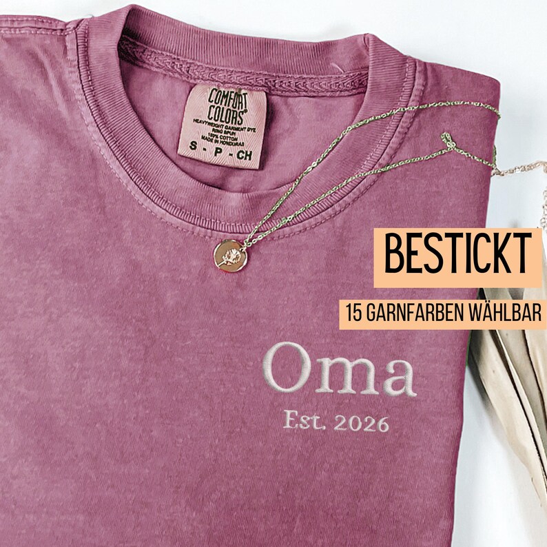 May include: A dusty rose t-shirt with the word "Oma" embroidered in white, along with "Est. 2026". A gold necklace with a pendant rests on the shirt. The shirt has a Comfort Colors tag and text that reads "BESTICKT" and "15 GARNFARBEN W&Auml;HLBAR".