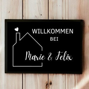May include: A black rectangular sign with white text and a house outline. The sign reads "WILLKOMMEN BEI Marie & Felix" with a heart above the house. The background is a wooden wall.