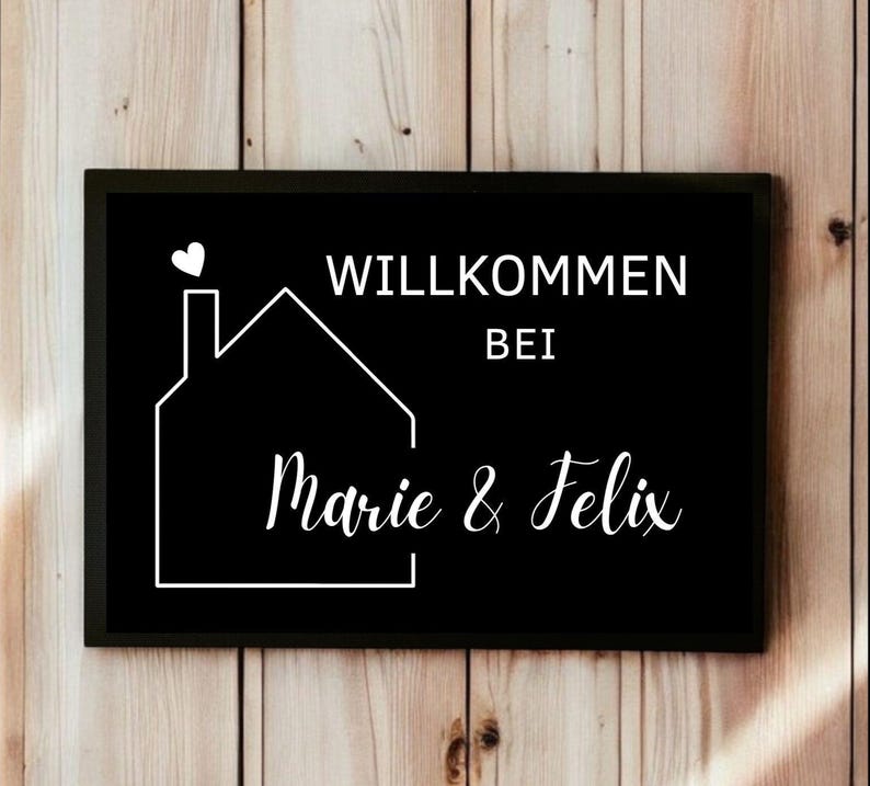 May include: A black rectangular sign with white text and a house outline. The sign reads "WILLKOMMEN BEI Marie & Felix" with a heart above the house. The background is a wooden wall.