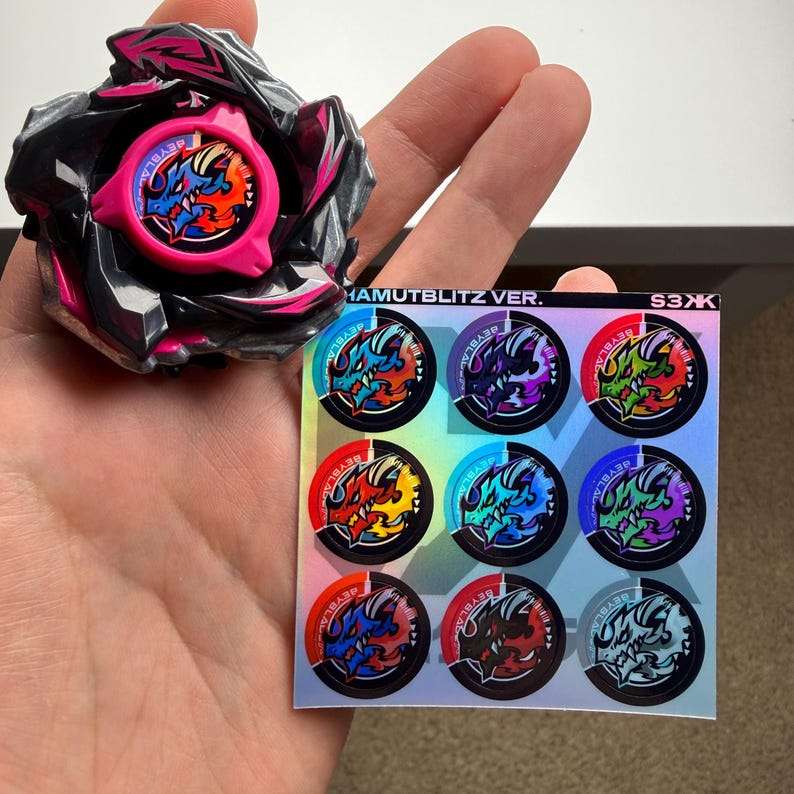 Puede incluir: Una peonza negra y rosa con un emblema de drag&oacute;n central, acompa&ntilde;ada de una hoja de pegatinas cuadrada con nueve dise&ntilde;os de dragones circulares en varios colores y el texto HAMUTBLITZ VER. S3XK impreso en la parte superior.