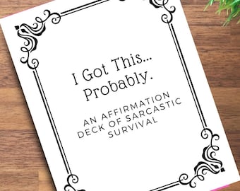 May include: A white card with black decorative borders and text that reads "I Got This... Probably. An Affirmation Deck of Sarcastic Survival." The card is on a wooden surface with a small green plant in the background.