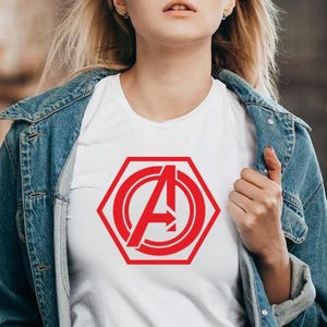 May include: White t-shirt featuring a red hexagonal design with the letter "A" and an arrow. The shirt is worn under a blue denim jacket. The design is a bold, graphic element.