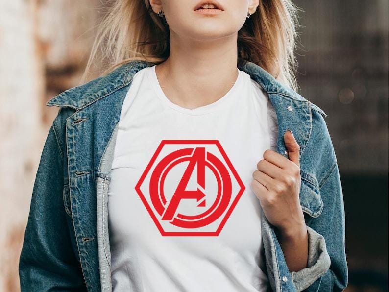 May include: White t-shirt featuring a red hexagonal design with the letter "A" and an arrow. The shirt is worn under a blue denim jacket. The design is a bold, graphic element.