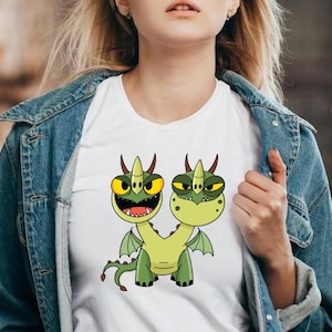 May include: White t-shirt featuring a cartoon illustration of a two-headed green dragon with yellow eyes, horns, and wings. The dragon is smiling with its mouth open. The shirt is being worn.