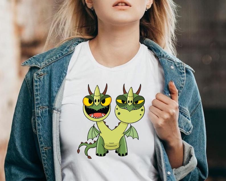 May include: White t-shirt featuring a cartoon illustration of a two-headed green dragon with yellow eyes, horns, and wings. The dragon is smiling with its mouth open. The shirt is being worn.