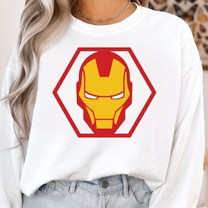 May include: White sweatshirt featuring a red and yellow Iron Man graphic. The graphic is a stylized depiction of Iron Man's helmet, enclosed within a red hexagon. The sweatshirt is a long-sleeved, casual top.