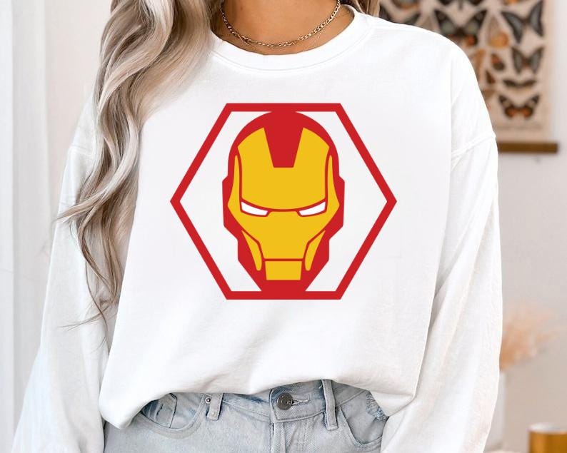 May include: White sweatshirt featuring a red and yellow Iron Man graphic. The graphic is a stylized depiction of Iron Man's helmet, enclosed within a red hexagon. The sweatshirt is a long-sleeved, casual top.