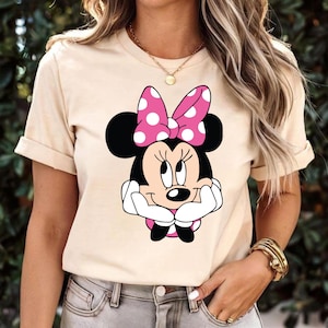 May include: Beige t-shirt featuring a Minnie Mouse graphic. The graphic shows Minnie Mouse with a pink polka-dot bow and white gloves. The shirt has short sleeves and a crew neck.