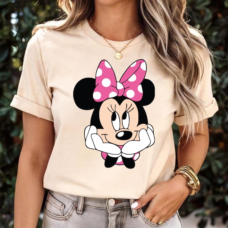 May include: Beige t-shirt featuring a Minnie Mouse graphic. The graphic shows Minnie Mouse with a pink polka-dot bow and white gloves. The shirt has short sleeves and a crew neck.