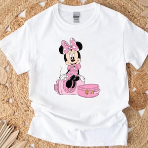May include: White t-shirt featuring a Minnie Mouse graphic. Minnie is depicted in a pink dress and bow, sitting on a pink suitcase. A smaller pink case is next to the suitcase. The design is centered on the shirt.
