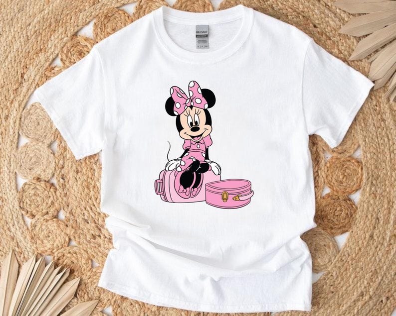 May include: White t-shirt featuring a Minnie Mouse graphic. Minnie is depicted in a pink dress and bow, sitting on a pink suitcase. A smaller pink case is next to the suitcase. The design is centered on the shirt.