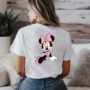 May include: White t-shirt featuring a Minnie Mouse graphic on the back. The cartoon character is depicted in a pink polka-dot dress and bow, with black ears and shoes. The shirt is a casual, everyday wear item.