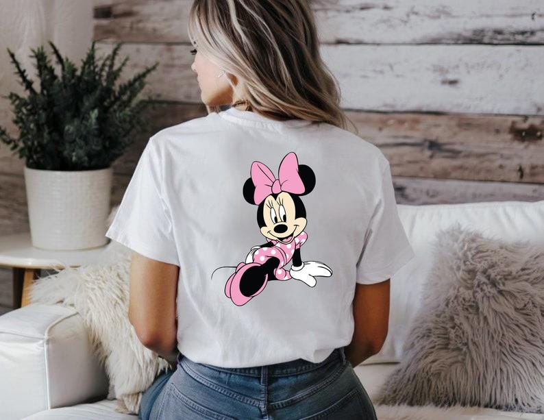May include: White t-shirt featuring a Minnie Mouse graphic on the back. The cartoon character is depicted in a pink polka-dot dress and bow, with black ears and shoes. The shirt is a casual, everyday wear item.