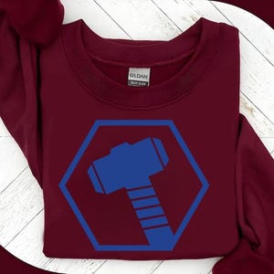 May include: A maroon sweatshirt featuring a blue hexagon with a blue hammer graphic. The sweatshirt has a crew neck and long sleeves. The brand name "Gildan" is visible on the tag.