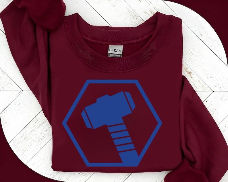 May include: A maroon sweatshirt featuring a blue hexagon with a blue hammer graphic. The sweatshirt has a crew neck and long sleeves. The brand name "Gildan" is visible on the tag.