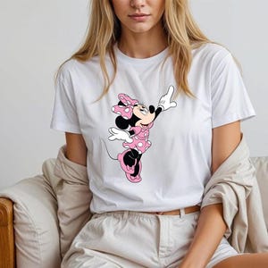 May include: White t-shirt featuring a graphic of Minnie Mouse in a pink polka-dot dress and bow. The cartoon character is in a playful pose with one arm raised. The shirt is worn by a person.