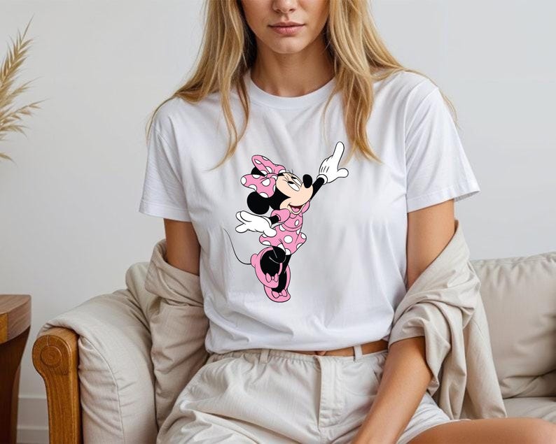 May include: White t-shirt featuring a graphic of Minnie Mouse in a pink polka-dot dress and bow. The cartoon character is in a playful pose with one arm raised. The shirt is worn by a person.