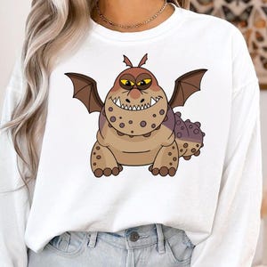 May include: White sweatshirt featuring a cartoon dragon with brown and purple spots, wings, and a toothy grin. The dragon has yellow eyes and is centered on the front of the shirt. The sweatshirt has long sleeves and a crew neck.