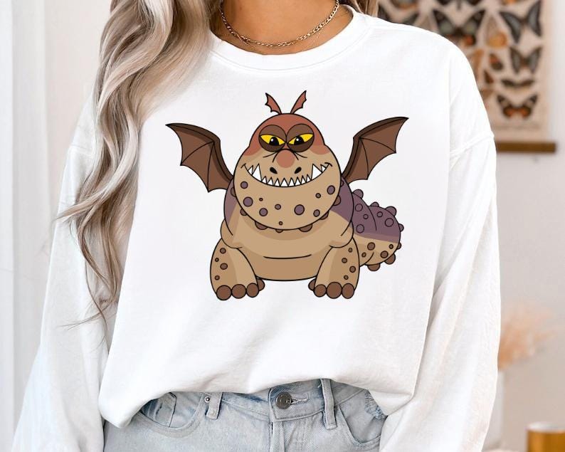 May include: White sweatshirt featuring a cartoon dragon with brown and purple spots, wings, and a toothy grin. The dragon has yellow eyes and is centered on the front of the shirt. The sweatshirt has long sleeves and a crew neck.