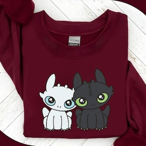 May include: A maroon sweatshirt featuring a cartoon graphic of two dragons. One dragon is white with blue eyes, and the other is black with green eyes. The sweatshirt has a crew neck and long sleeves.