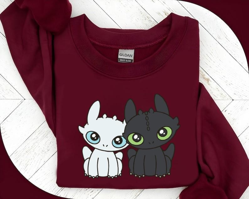 May include: A maroon sweatshirt featuring a cartoon graphic of two dragons. One dragon is white with blue eyes, and the other is black with green eyes. The sweatshirt has a crew neck and long sleeves.