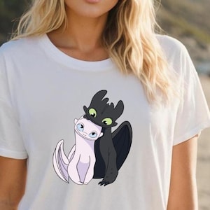 May include: White t-shirt featuring a graphic of two cartoon dragons, one black and one white, embracing. The black dragon has green eyes and the white dragon has blue eyes. The shirt is made of a soft, light-colored material.