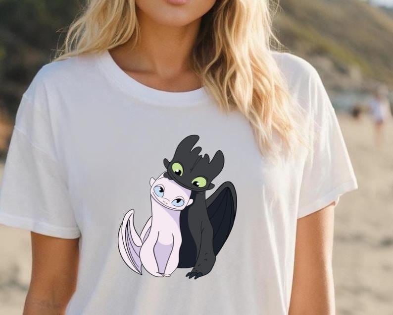 May include: White t-shirt featuring a graphic of two cartoon dragons, one black and one white, embracing. The black dragon has green eyes and the white dragon has blue eyes. The shirt is made of a soft, light-colored material.