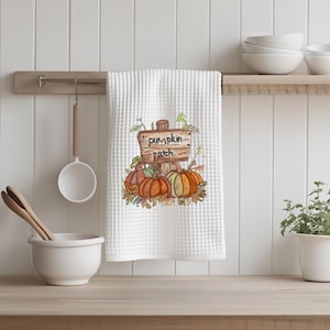 May include: White waffle-weave kitchen towel with a watercolor illustration of pumpkins and a wooden sign that reads "pumpkin patch." The pumpkins are orange, green, and yellow. The towel is hanging on a wooden bar.