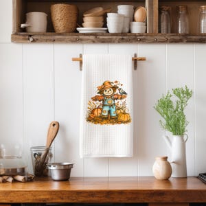 May include: White waffle-weave kitchen towel featuring a colorful illustration of a scarecrow with a hat, overalls, and pumpkins. The towel is hanging on a wooden rod in a kitchen setting. The design has autumn colors.