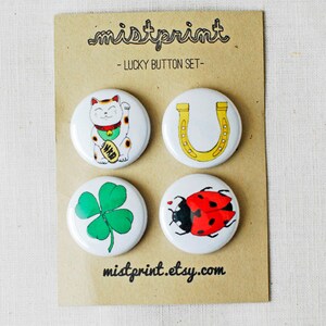 Può includere: Set di quattro bottoni con ciondoli portafortuna. I bottoni sono bianchi con un contorno nero e presentano un gatto fortunato, un ferro di cavallo, un quadrifoglio e una coccinella.