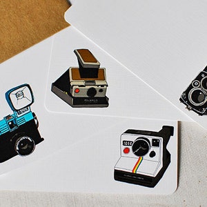 May include: Four vintage cameras on white cards. The cameras are a blue and black Polaroid, a black and brown Polaroid, a black and white Polaroid, and a black and silver film camera.