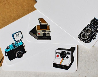 Pode incluir: Quatro câmeras vintage em cartões brancos. As câmeras são uma Polaroid azul e preta, uma Polaroid preta e marrom, uma Polaroid preta e branca e uma câmera de filme preta e prateada.