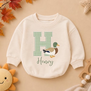 May include: A cream-colored long-sleeve sweatshirt featuring a large green and white gingham letter H. Next to the letter is a graphic of a mallard duck with a green head, brown chest, and white body. The name Henry is printed in green script below.