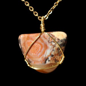 May include: A pendant necklace featuring a triangular, patterned stone in shades of orange, brown, and white. The stone is wrapped in gold-colored wire and suspended from a delicate gold chain. The background is solid black.