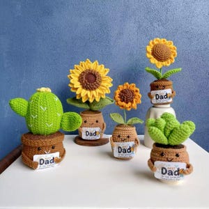 Pode incluir: Uma coleção de plantas de crochê feitas à mão, incluindo girassóis, um cacto e uma suculenta, cada uma em um vaso marrom. Os vasos têm rostos sorridentes e seguram placas que dizem "Best Dad". As flores são amarelas e marrons.