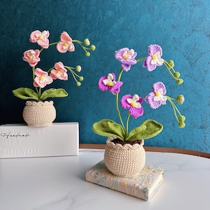 Puede incluir: Dos plantas de orquídeas de ganchillo en macetas beige. Una tiene flores rosas, la otra flores moradas. Se ven las hojas y los tallos verdes. Las macetas están sobre una superficie blanca y un libro. El texto "Handmade GIFT FOR YOU" está en la superficie blanca.