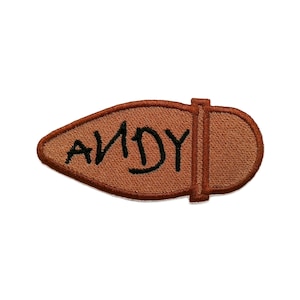 Op de afbeelding: Een bruine geborduurde patch in de vorm van een schoenzool met een donkerbruine rand. Het woord ANDY is met zwarte draad in het midden van de patch geborduurd. Het ontwerp heeft een verticale stiklijn die de hiel van de teen scheidt.