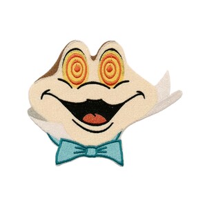 May include: An embroidered patch featuring a cartoon frog face with cream-colored skin, a wide black mouth, and orange spiral eyes. The frog wears a light blue bow tie and has a white collar-like detail extending from the sides of its head.