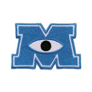 May include: A blue embroidered patch in the shape of the letter M, featuring a white and black eye graphic centered within the letter. The patch has a thick white border outlining the blue letter shape, creating a distinct and bold visual design.