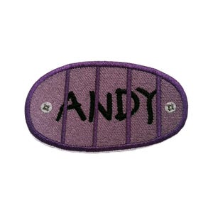 May include: An oval-shaped embroidered patch featuring a light purple background with vertical dark purple stripes. The name ANDY is stitched in black capital letters across the center. Two small white circular accents are on the left and right.