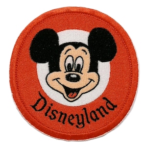 May include: Embroidered patch featuring Mickey Mouse's smiling face within a white circle, set against a vibrant orange background. The word "Disneyland" is arched below the character in black script. The patch has a stitched border.