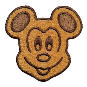 May include: Embroidered patch of a smiling cartoon mouse face with large ears. The patch has a tan background with brown outlining and details. The mouse has large oval eyes and a wide smile. Suitable for clothing or accessories.