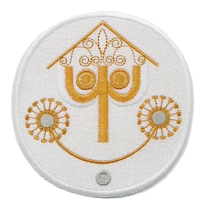May include: Embroidered patch featuring a stylized face with a house-like top and floral accents. The design is primarily gold on a white background, with a circular shape and a small gray detail at the bottom.