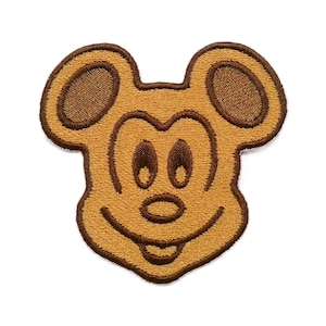 May include: An embroidered patch featuring the iconic head shape of a cartoon mouse. The design is rendered in shades of golden tan and dark brown, with stitched outlines defining the ears, eyes, nose, and a wide, smiling mouth.