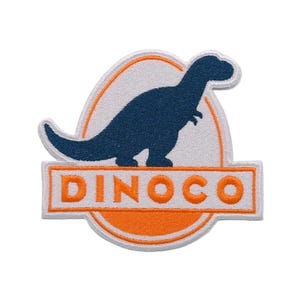 May include: An embroidered patch featuring a dark blue silhouette of a dinosaur standing inside an oval frame with an orange border. Below the dinosaur, a rectangular orange banner contains the white text DINOCO in a bold, sans-serif font.