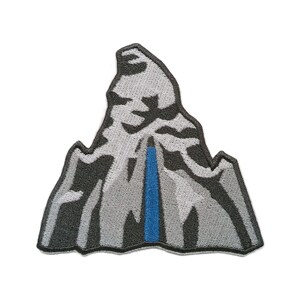 Può includere: Una toppa ricamata a forma di vetta di montagna con una silhouette frastagliata. Il design presenta sfumature di grigio chiaro, grigio scuro e nero per raffigurare texture rocciose e neve, con una striscia verticale blu brillante al centro.